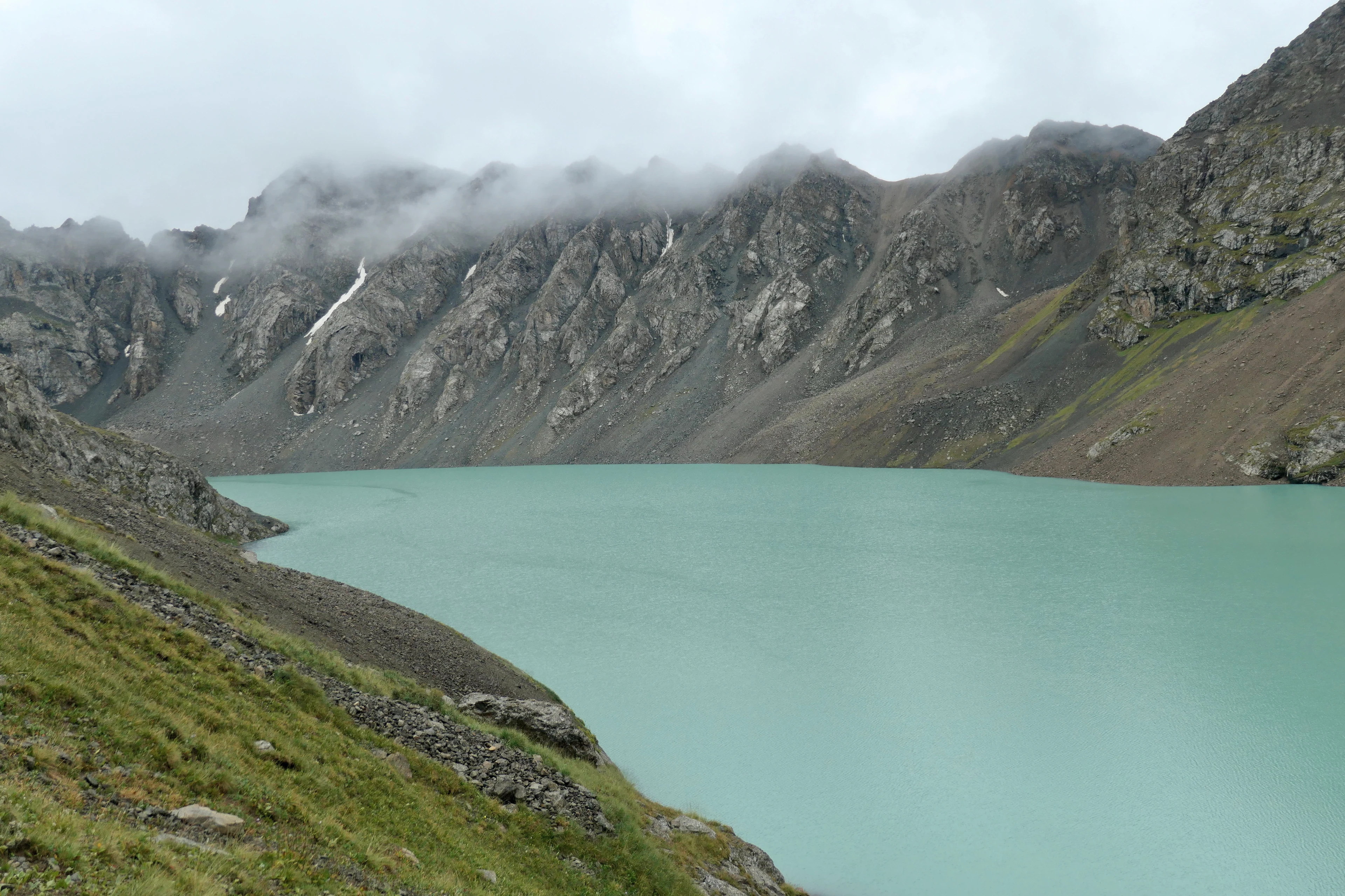 Turquoise Ala-Kul lake beneath rocky peaks and low cloud.