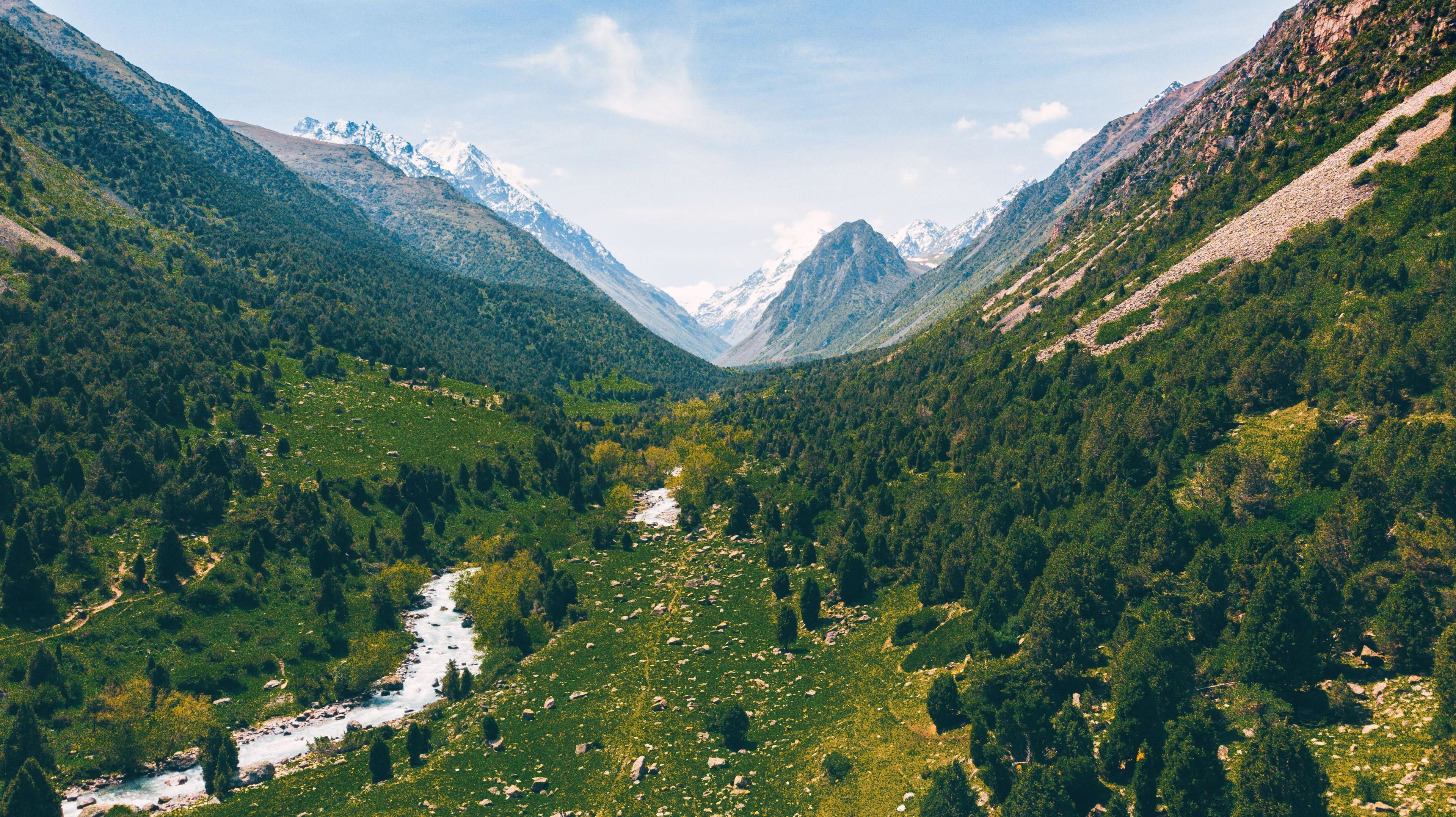 A river winds through forested slopes beneath the snowy mountains of Kyrgyzstan.